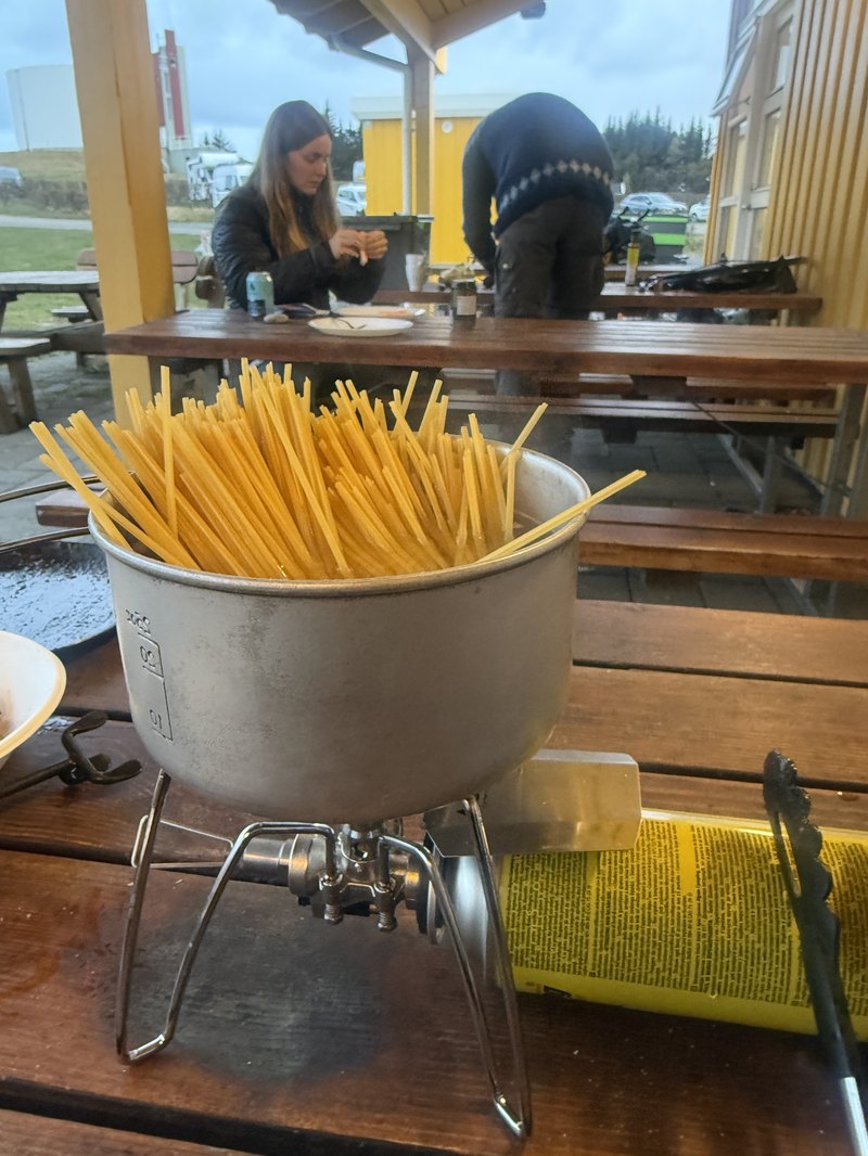 キャンプ場の屋根の下、携帯コンロの大鍋でパスタを茹でる様子と仲間たち